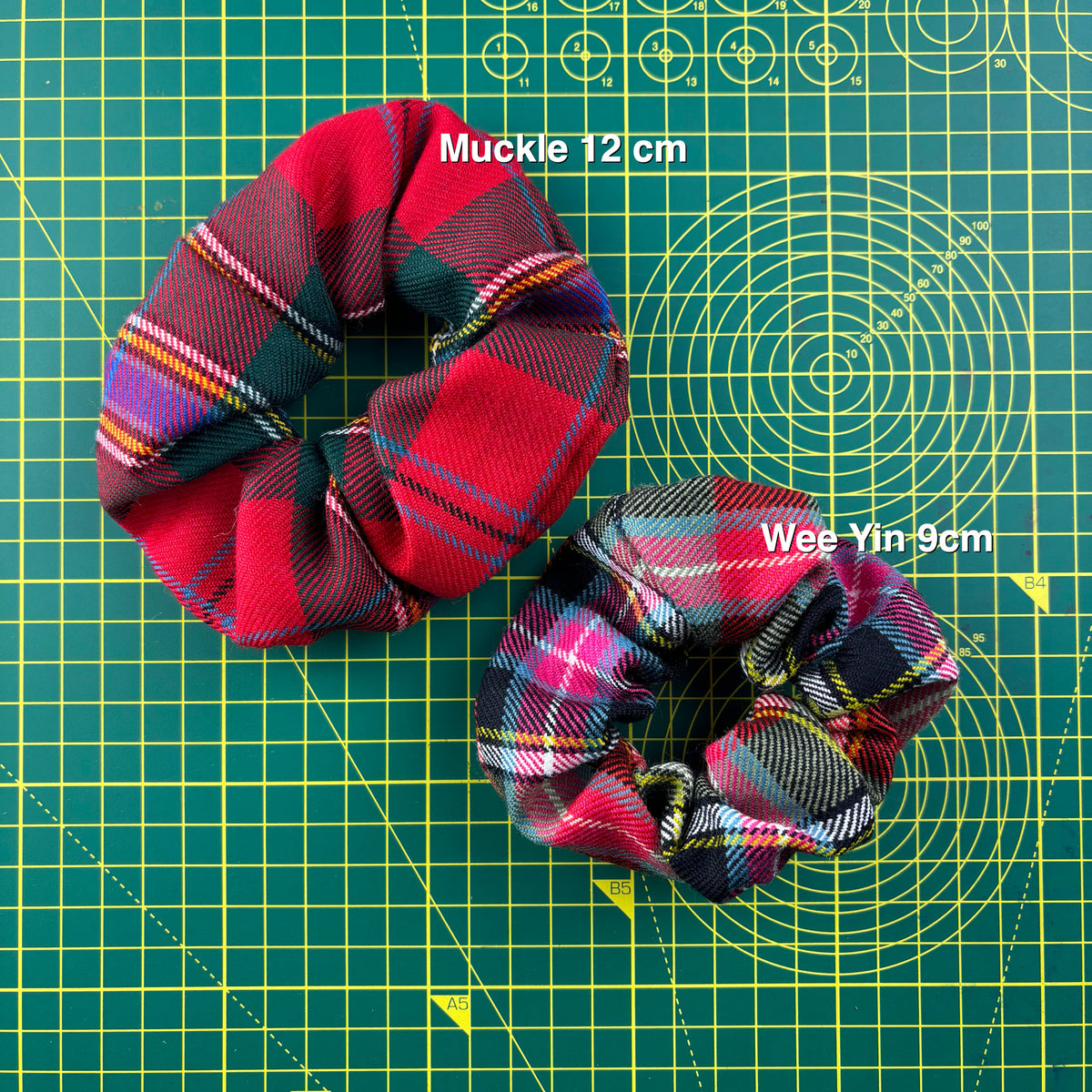 Two tartan scrunchies on a cutting mat showing the sizes: the larger Muckle (12 cm) and the smaller Wee Yin (9 cm).
